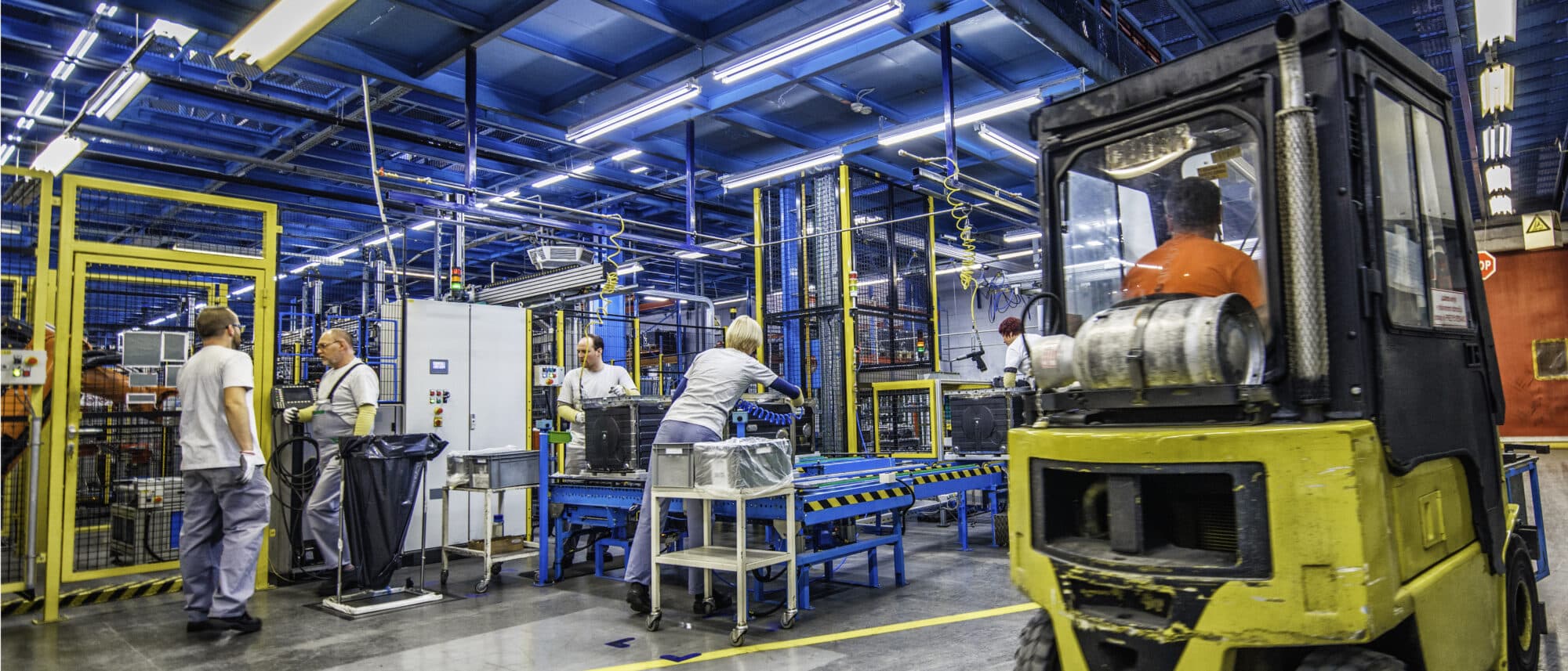 Factory workers doing their jobs while a forklift is driving by.